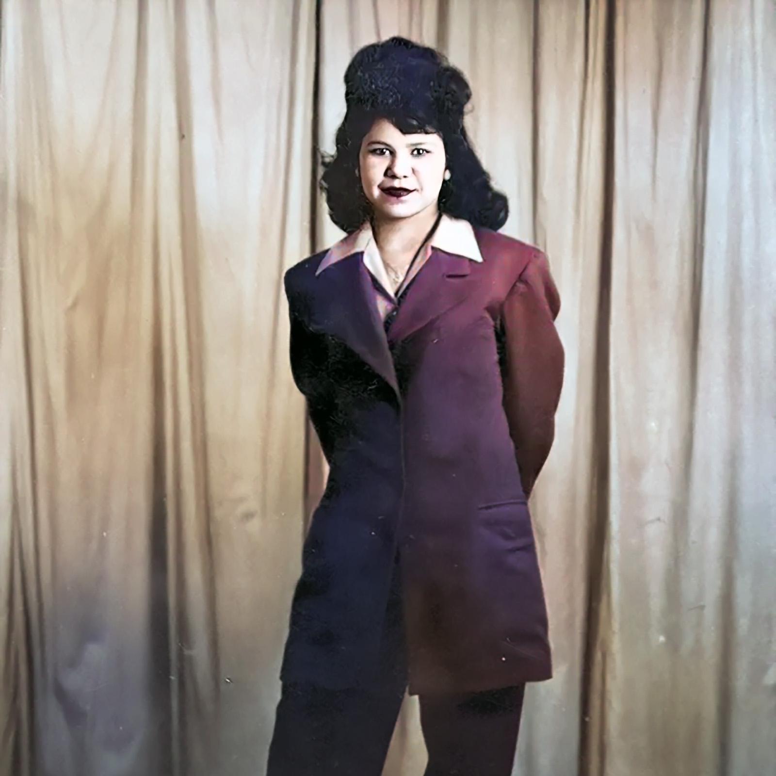 A young woman in a suit jacket and pants stands in front of a curtain. She has long black hair.
