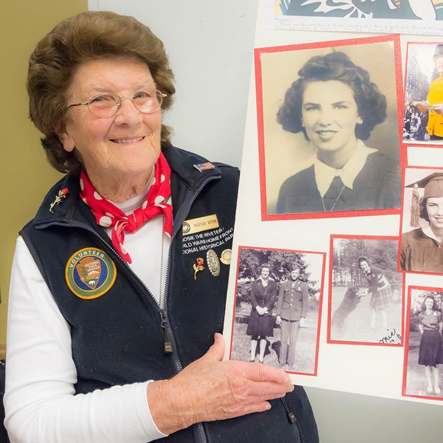 Older woman with curly hair and glasses holds a homemade poster with photos of herself.
