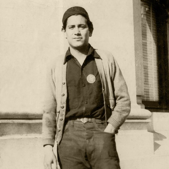 A young latin male in a hat, pants, shirts and a jacket is posing for the camera. 