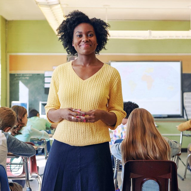 A female African American teachers smiles at the camera from the back of the classroom. 