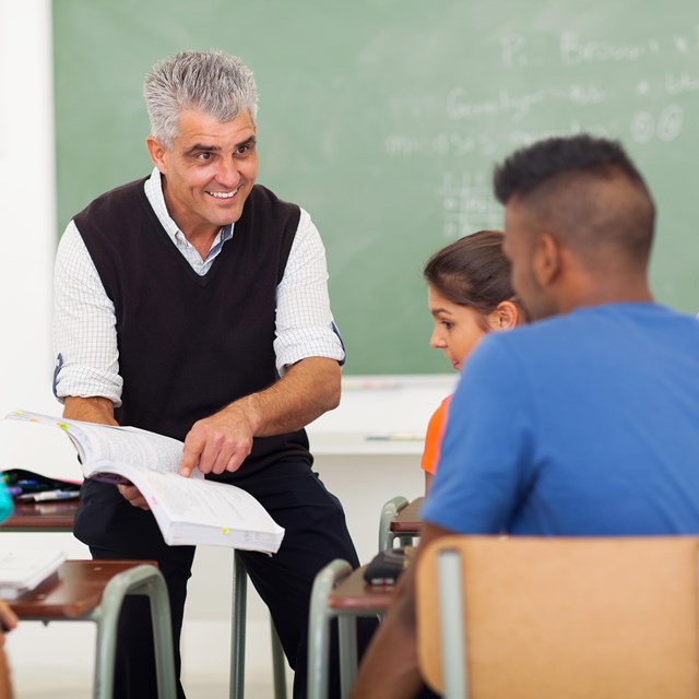 An older male teacher points to a book while teaching students. 