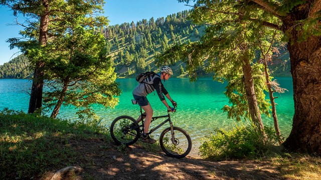 A biker rides down a dirt trail with trees alongside a clear water river on a sunny day. 