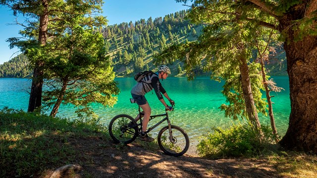 A biker rides down a dirt trail with trees alongside a clear water river on a sunny day.