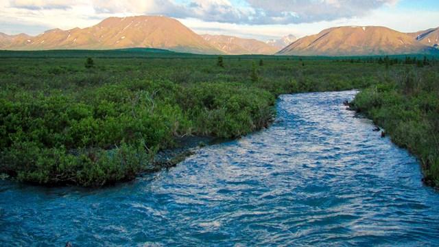 A blue river flows through greenery with distant hills and a blue cloud-filled sky. 