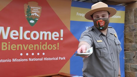 Park Ranger wearing a mask stands in front of welcome banner and holds a brochure toward the camera.