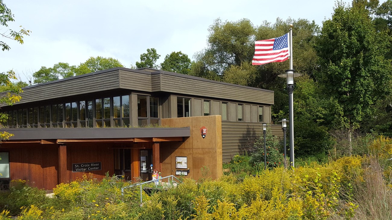 A brown two-story building surrounded by green vegetation. A flag on a flag pole waves in the wind.