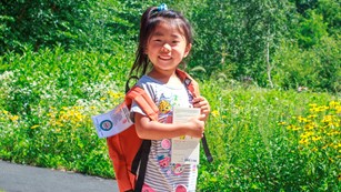 a girl wearing a backpack and holding a book stands near a garden