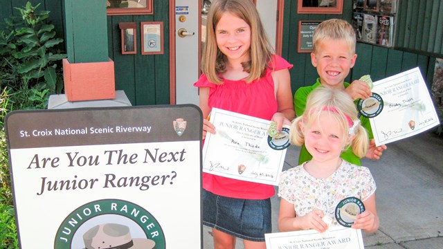 Three children hold certificates, badges, and patches from the Junior Ranger Program