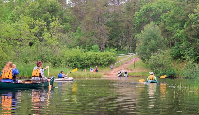 People canoeing and kayaking towards shore where other people are at the sandy and green landing