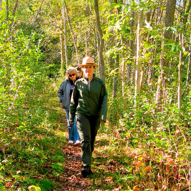 A park ranger and people walk along a wooded trail.