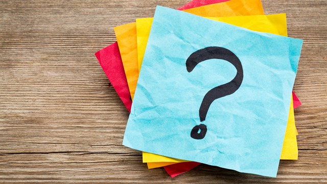 An image of four various colored post-it notes with a large question mark laid on top of a wood.