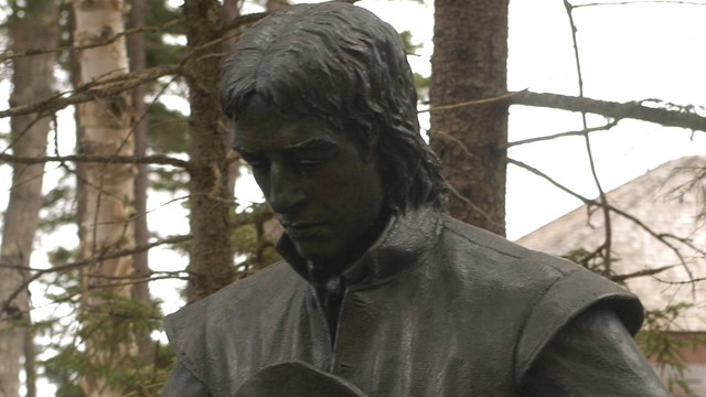 A bronze statue depicts a man in a long-sleeve shirt holding a broad-rimmed hat & looking down.