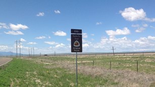 A road runs through a vast, flat grassland. A brown sign stands next to the road.