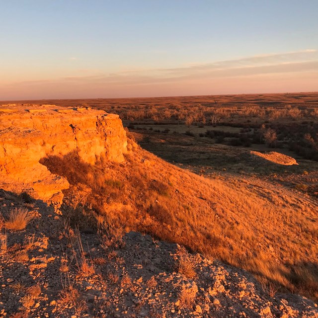 A rocky bluff overlooks the prarie, under a setting sun and warm light.