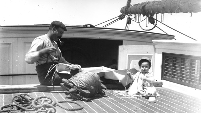Man smoking a pipe while holding rope with a small child beside him on the deck of a ship