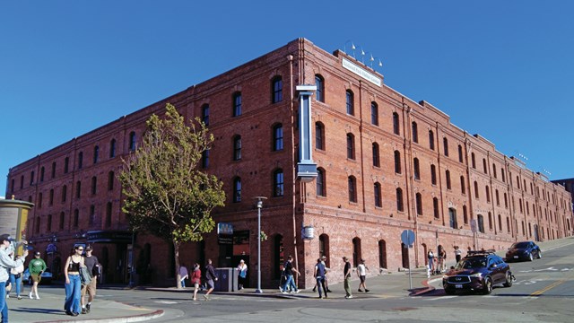 A historical brick warehouse stands on the corner of a busy street on a sunny day.