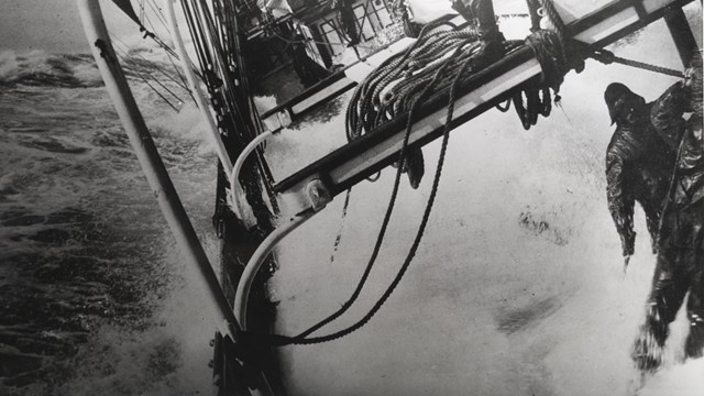 Ocean waves crashing onto the deck of a ship with two men hanging on to the ship