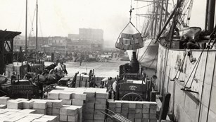 Boxes of cargo stacked on a pier beside a ship, a sling attached to a mast lifting cargo aboard