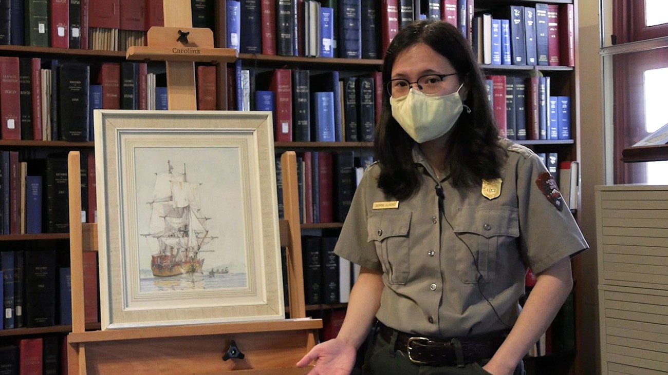Ranger standing beside a painting of a ship on an easel with bookshelves in the background