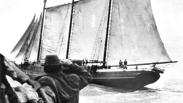 Black and white photo of a man on shore waving to a sailing ship on the water.