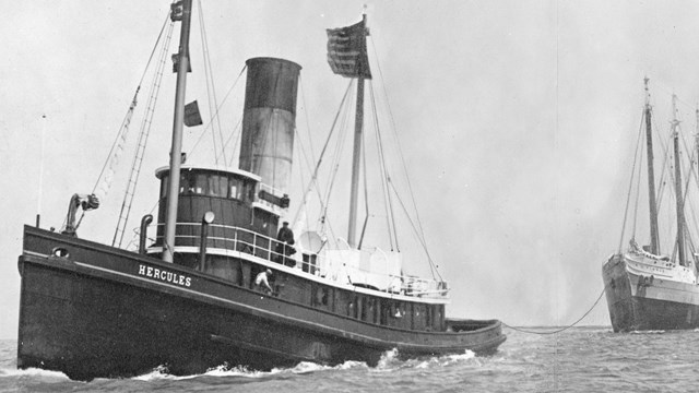 Black and white photo of a tugboat towing a sailing ship on the ocean,