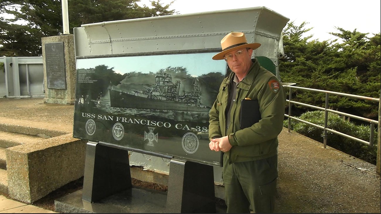 Park ranger standing outside beside a wayside exhibit showing the image of a ship