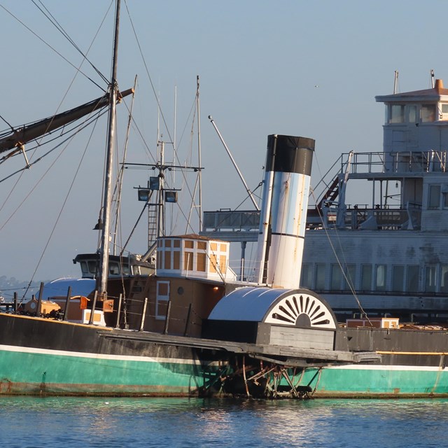 a medium-sized steamship with large side wheels and a green hull is docked to a pier