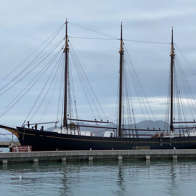A large black wooden ship with three masts is docked to a pier