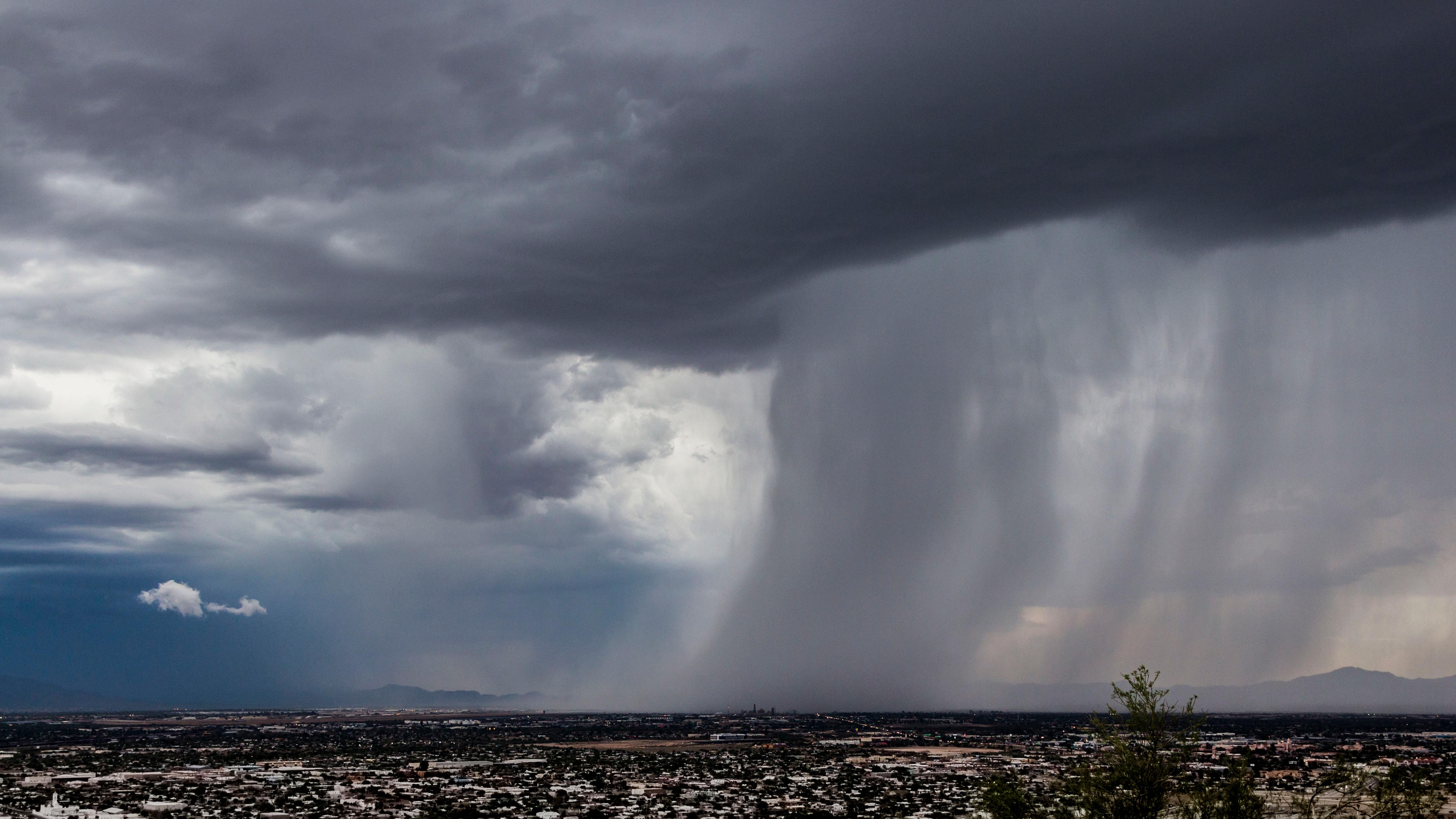 A large storm cloud moves left, over the city below. Pouring rain throughout the right of the image