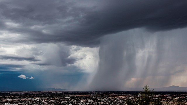 A large storm cloud moves left, over the city below. Pouring rain throughout the right of the image