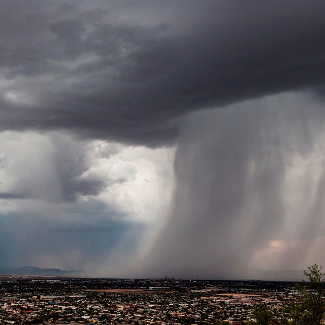 Image looks out over the city of Tucson. A large storm cloud rolls in from the right, dumping water.