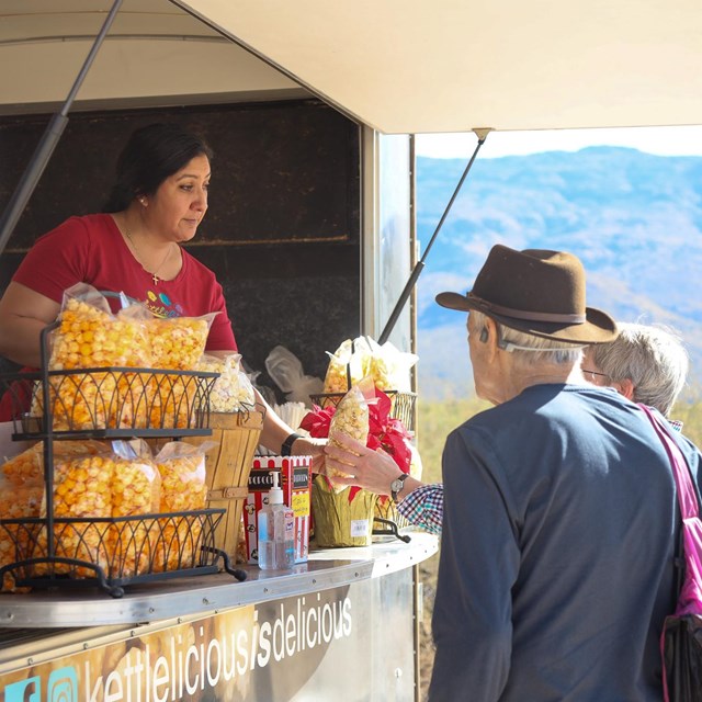 An older couple stand (right) look into a food truck (left) where a women hands them popcorn.