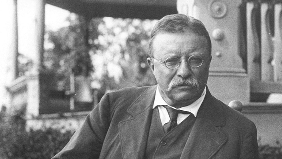 A black and white photo of Theodore Roosevelt seated in front of the porch of Sagamore Hill.