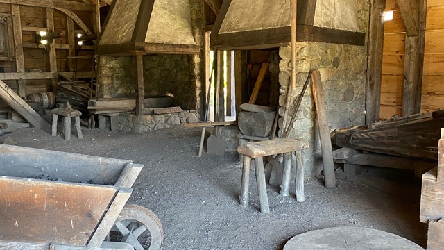Building interior with two chimneys, gravel floor, stool, wheelbarrow, and other blacksmith tools