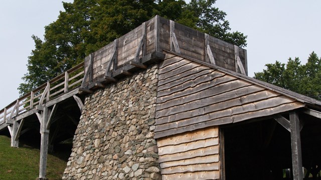 A wooden bridge leads to a large, stone furnace with a wooden shed attached.
