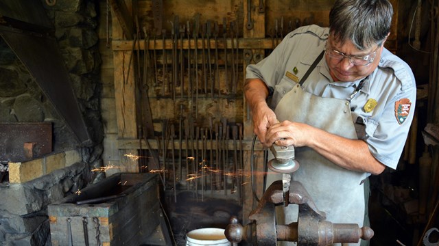 Park ranger uses a metal grinder on a piece of metal while in a shop lined with tools.