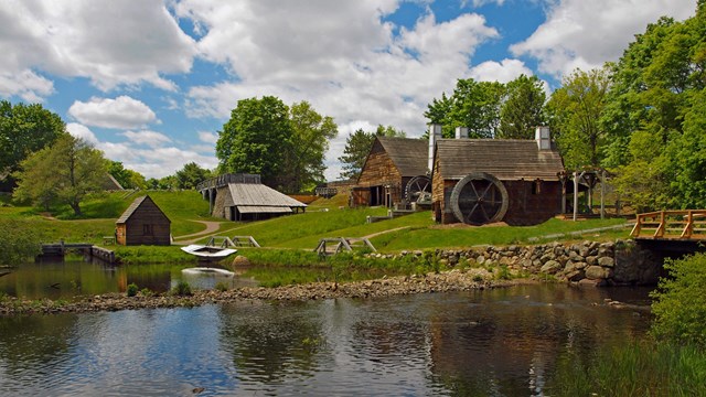 Multiple historic wooden structures line a river bank shaded by green trees.