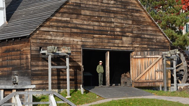 Ranger stands in doorway of a large wood-sided structure used to forge iron.