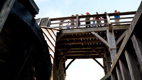 Group of people stand on bridge looking down on waterwheel.