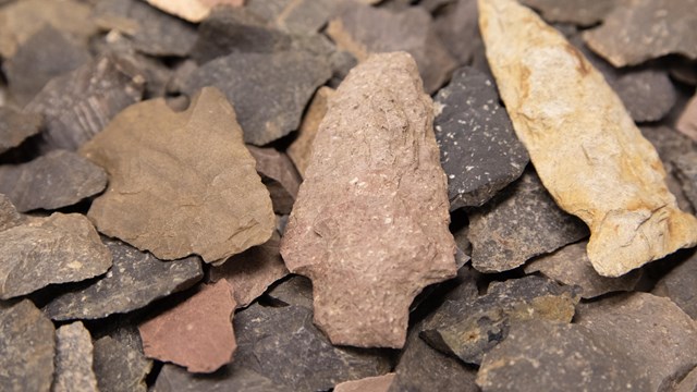A pile of different size, shape, and color chips of stone with five projectile points on top.