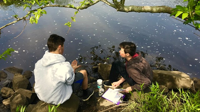 Two students sit by a river. 