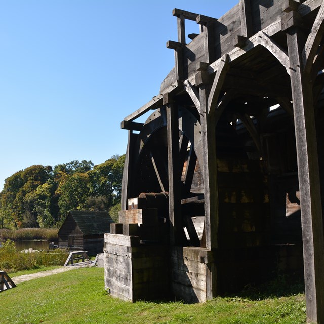 wooden structure with large wheel on a hill overlooking a river under a blue sky.