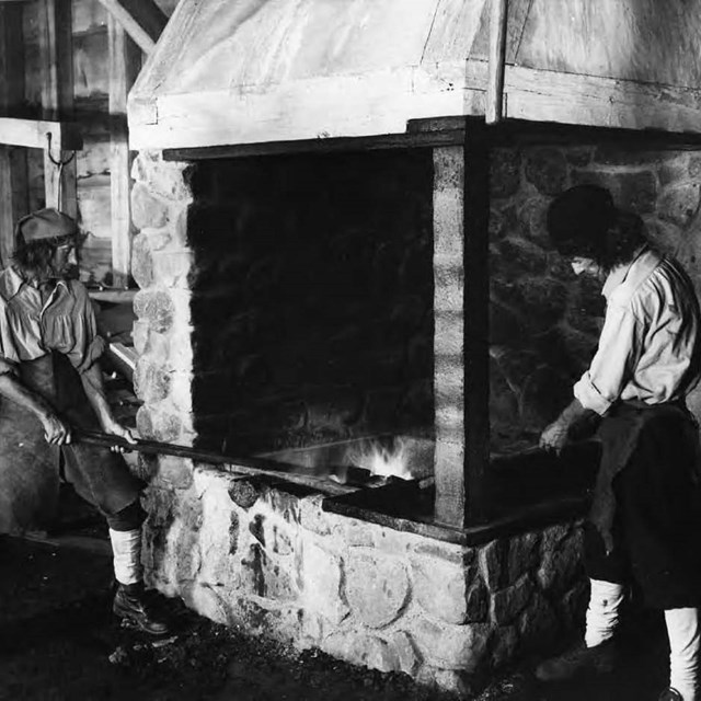 Two men in white shirts and dark hats standing around a fire.