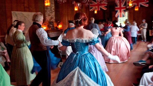 Volunteers dressed in period clothing dance in a historic building.