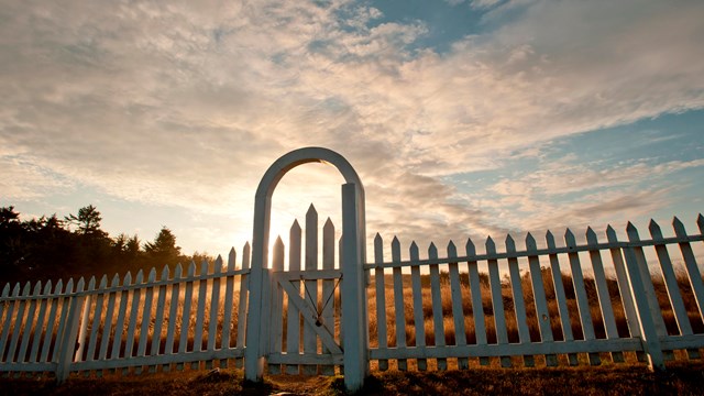The sun rises over a white fence.