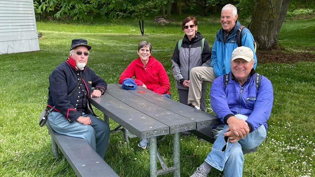 Five older white people sit and stand around a picnic table and bench