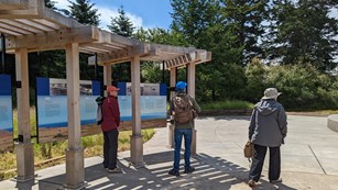 Three visitors stand outside of the new visitors center. Educational signs in the background