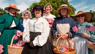 Volunteers dressed in historic garb smile while holding American flags.