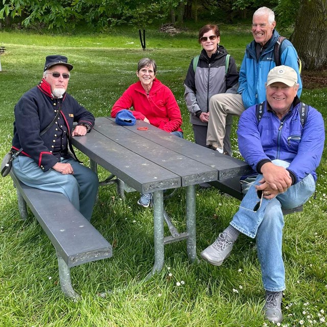 Five older white people sit and stand around a picnic table and bench