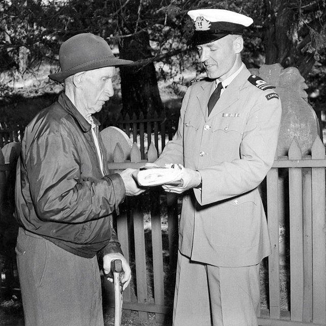 Black and white photo. A man in military uniform hands another man a package.